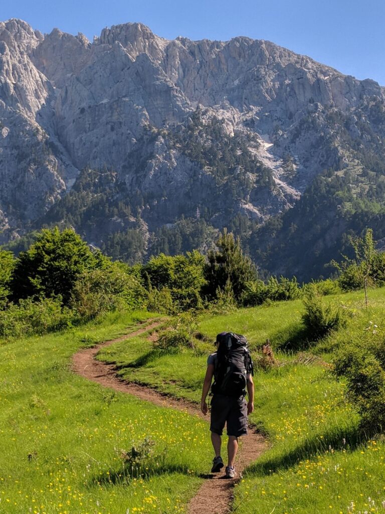 A hiker follows a grassy trail beneath the rugged mountains of the Valbona to Theth route in Albania