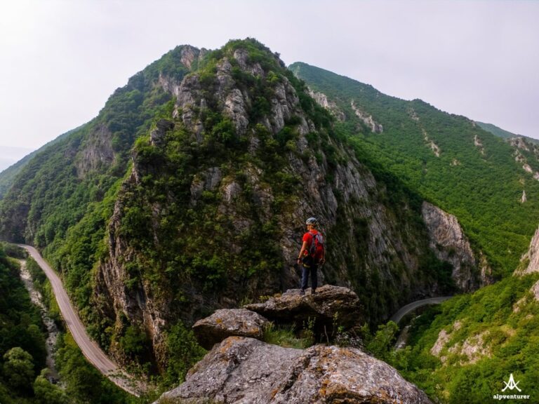 A hiker in red stands on a rocky ledge above a winding road and green cliffs at Via Ferrata Prizren