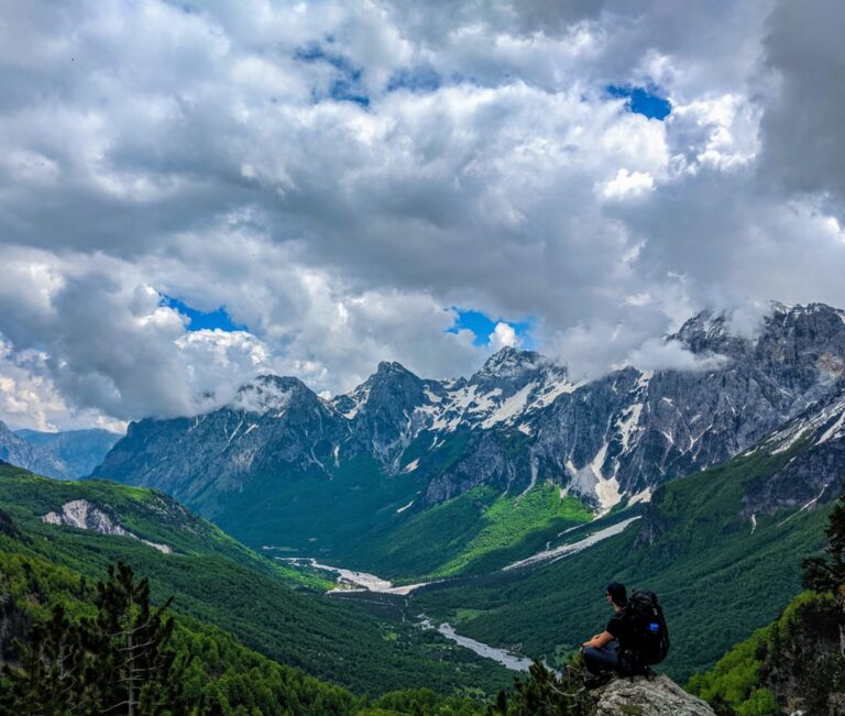 A hiker overlooks the green Valbona Valley and rugged peaks beneath a cloudy sky