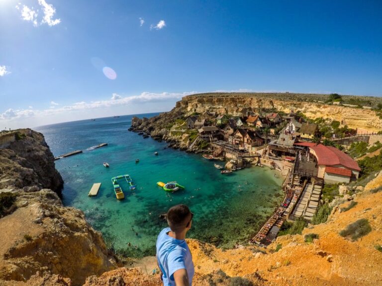 A hiker overlooks a turquoise cove and rocky cliffs on Gozo under a bright blue sky