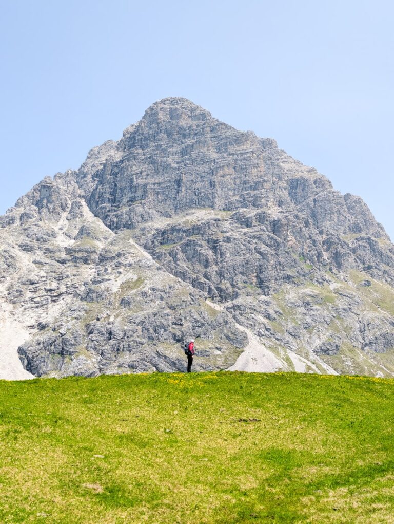 A hiker stands on a green slope below the rocky summit of Großer Widderstein under a clear sky