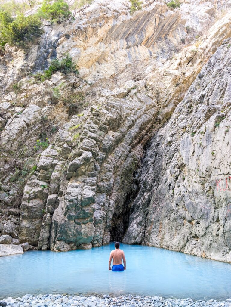 A hiker stands in turquoise water beneath the steep pale rock walls of Holta Canyon