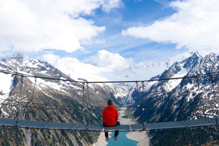 A hiker in a red jacket sits on the Olpererhütte suspension bridge above a snowy alpine valley