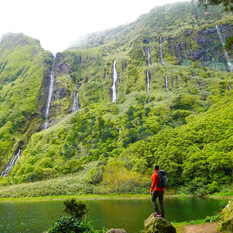 A hiker stands beside a pool beneath the green waterfall covered cliffs of Poço da Ribeira do Ferreiro on Flores