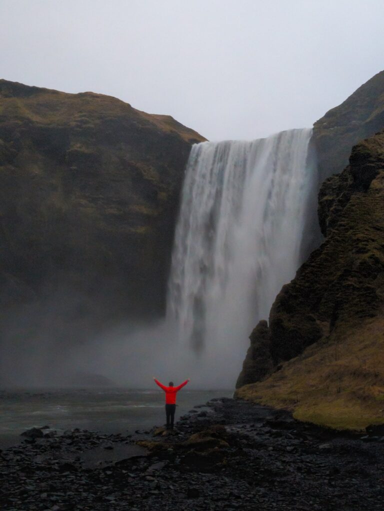 A hiker in a red jacket faces Skógafoss, framed by dark cliffs beneath an overcast sky