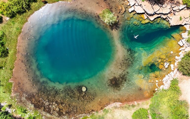 Aerial view of Syri i Sheganit’s deep blue karst spring ringed by shallow water and rocky shore