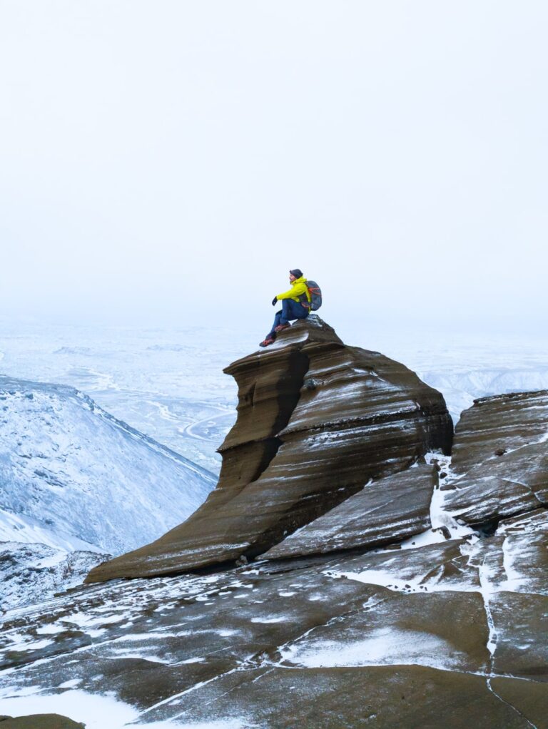 A hiker in a yellow jacket sits atop a layered rock outcrop on snow covered Vífilfell