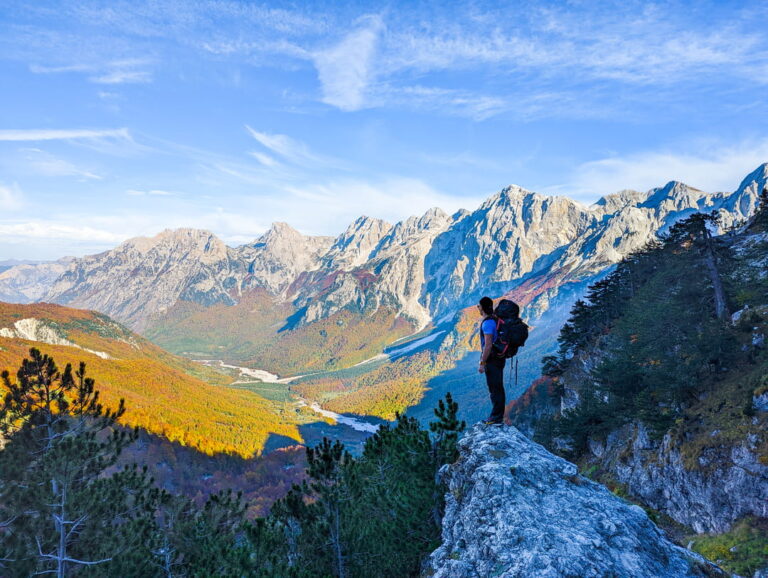 A hiker overlooks an autumn valley and rugged Albanian mountain peaks beneath a clear blue sky