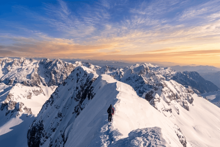 A hiker crosses a snowy ridge above the Jezerce mountains beneath a pink and blue evening sky