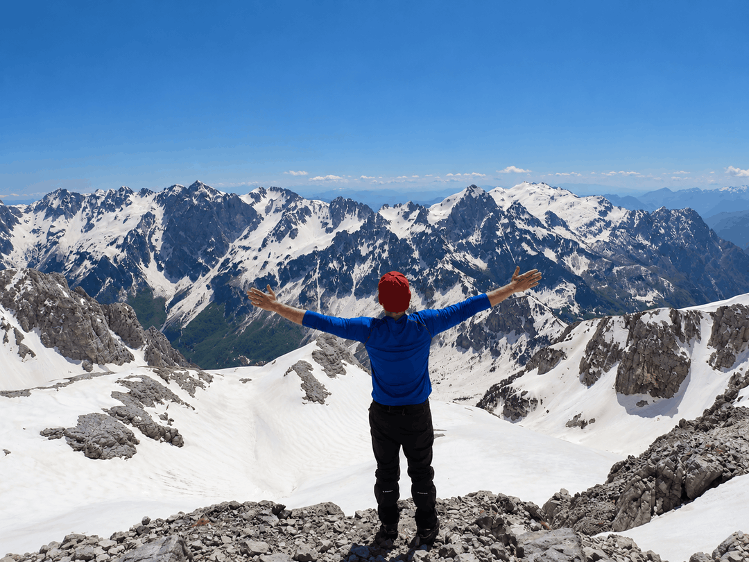 A hiker stands on a rocky, snow covered summit of Maja e Poplukës, facing a range of snow capped Albanian peaks