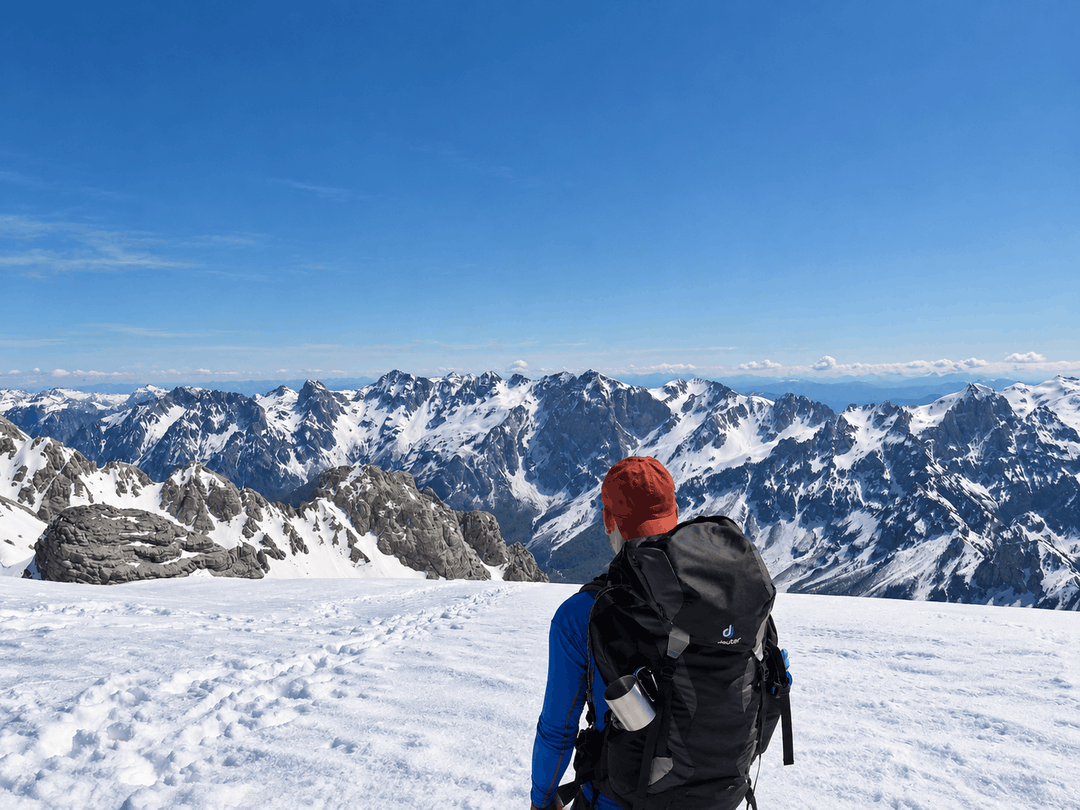 A hiker with a backpack looks across snow covered peaks from Maja e Poplukës in bright winter sunlight