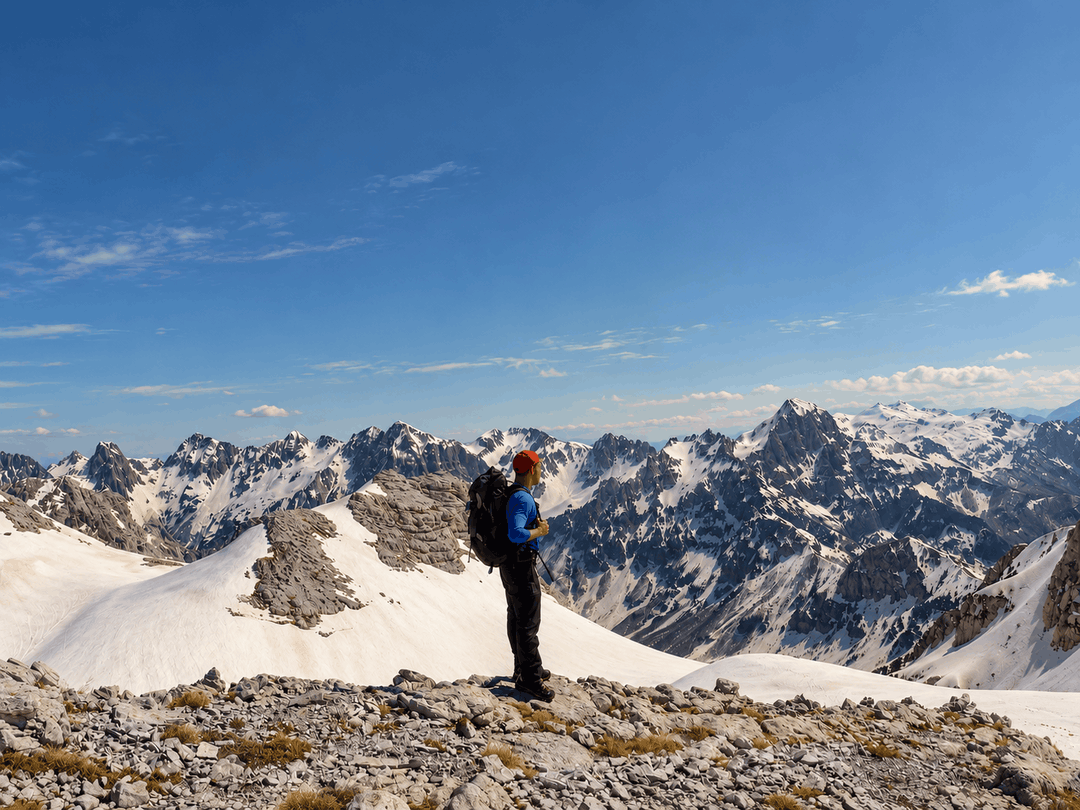A hiker on a rocky, snow-dusted ridge overlooking the Maja e Poplukës mountains beneath a clear blue sky