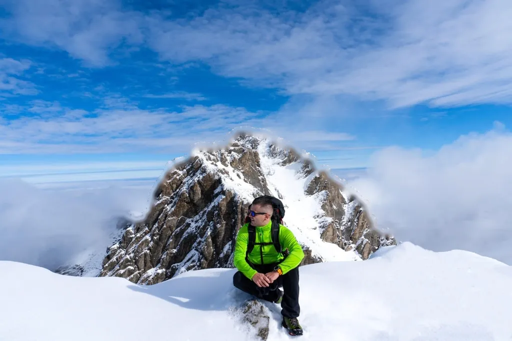 A hiker in a green jacket sitting in the snow below a rocky summit of Mount Olympus