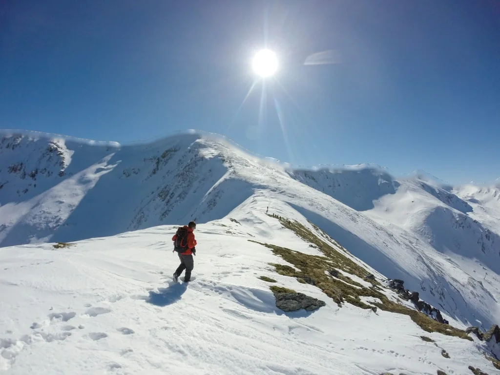 A hiker in red walking down a snowy ridge on Piribreg, the Sharr mountains behind