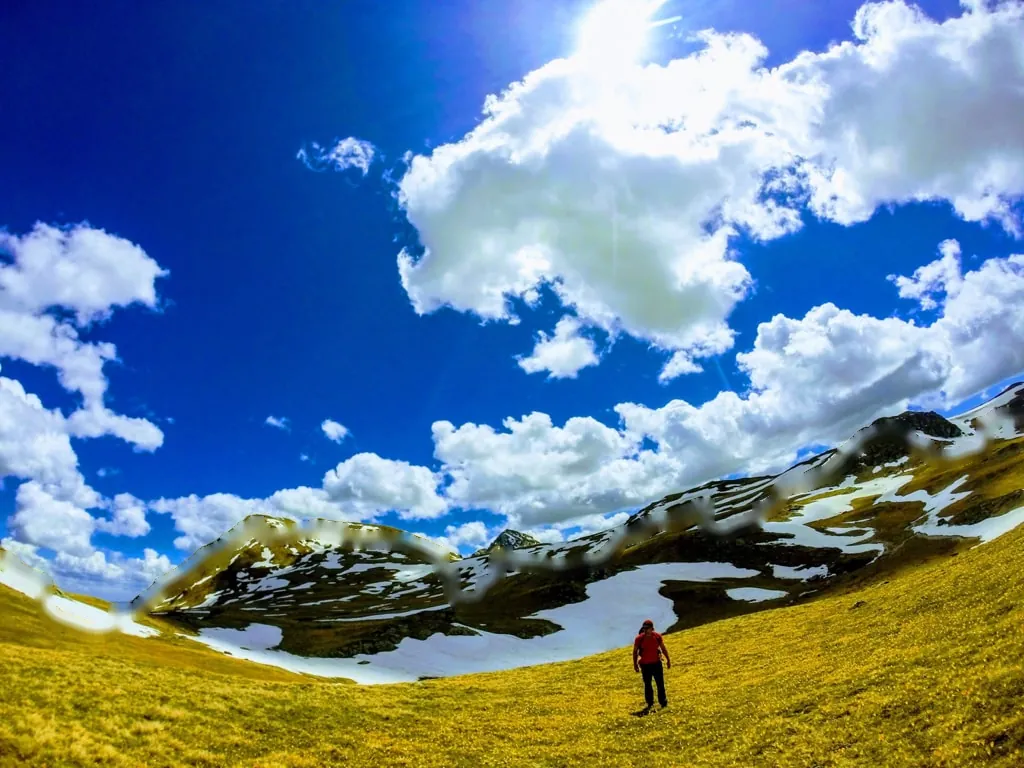 A hiker in red crossing a high meadow below the snow-patched ridge of Great Rudoka