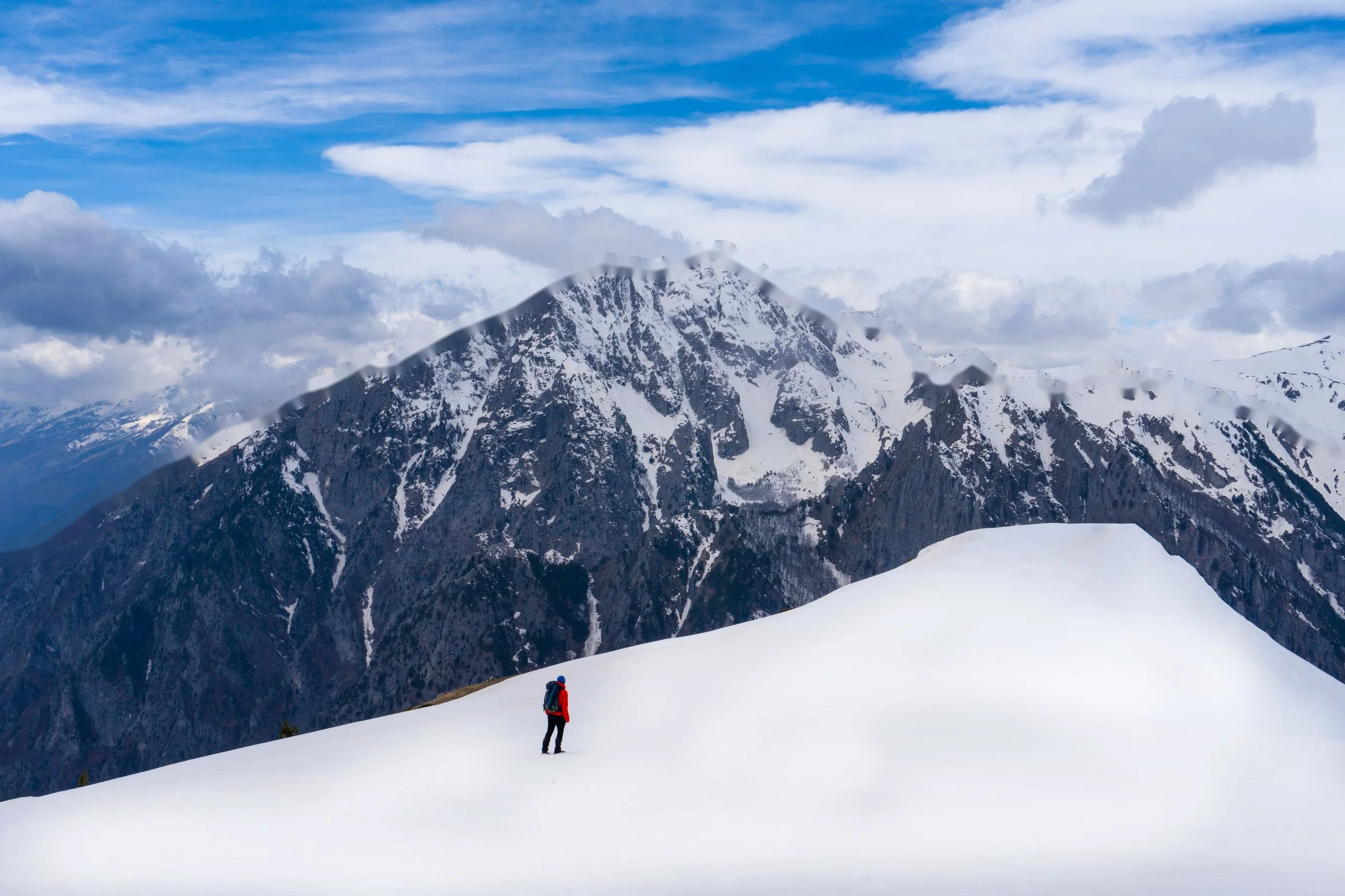 Ein Wanderer auf einem verschneiten Grat unterhalb des schneebedeckten Gipfels des Shkëlzen, darüber Wolken